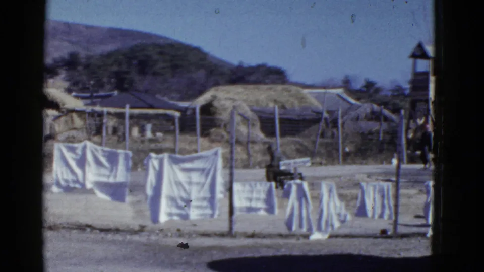 Rural village frame showing laundry on a fence, thatched huts, mountains, and a small tower-like structure at the right edge.