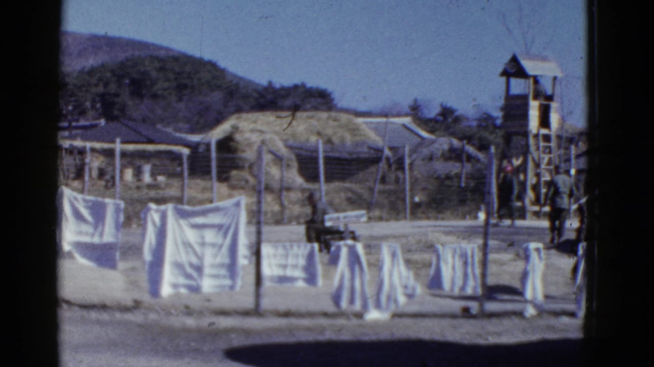 1950s San Francisco home-movie frame showing white laundry, fencing, a wooden guard tower, barracks, and uniformed men in a dusty compound.