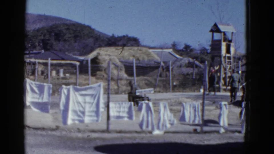 1950s San Francisco home-movie frame showing white laundry, fencing, a wooden guard tower, barracks, and uniformed men in a dusty compound.