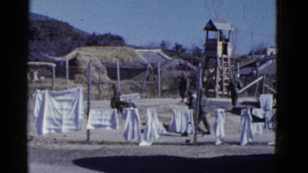 1950s San Francisco footage frame with soldiers moving through a fenced encampment behind white laundry and a wooden guard tower.