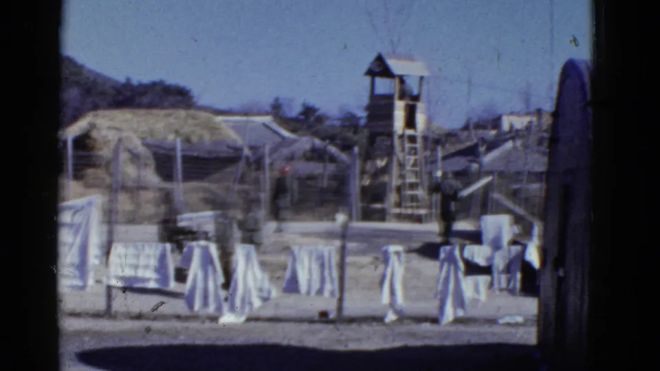 1950s San Francisco home-movie frame showing laundry, fencing, a guard tower, uniformed figures, and simple camp structures.
