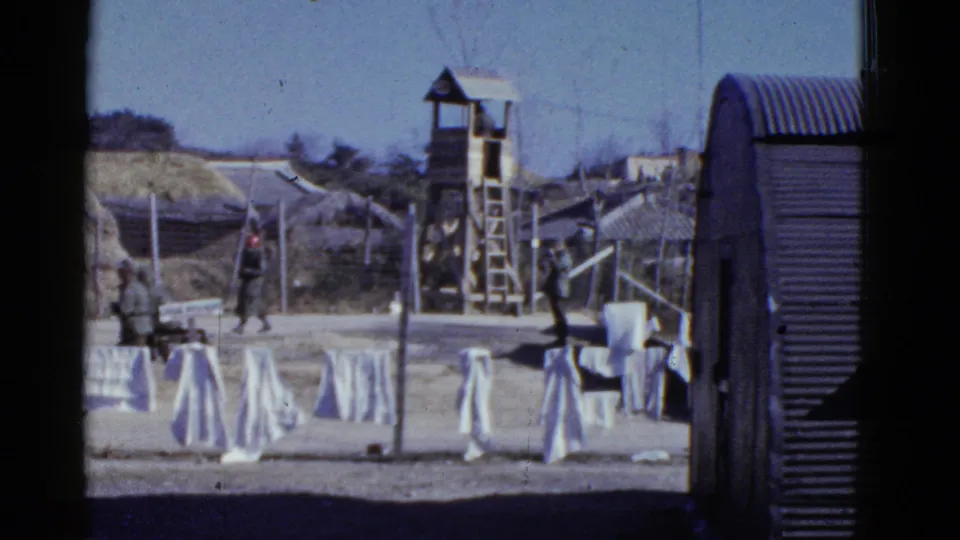1950s San Francisco frame with soldiers crossing a fenced encampment beside laundry lines, a wooden guard tower, and a curved metal hut.