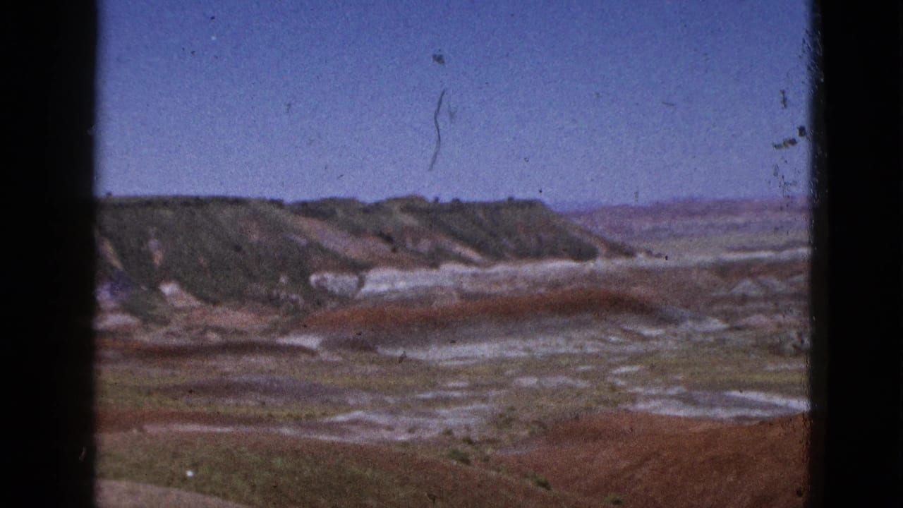 California desert scenery with distant eroded ridges, red-brown flats, pale mineral deposits, and a clear blue sky on 8mm film.