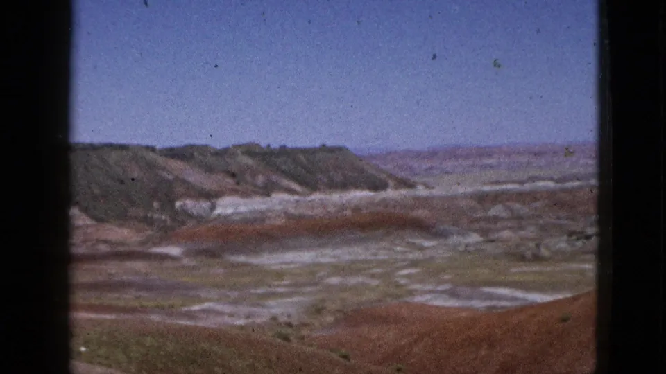 Wide 1950s 8mm view of a California desert basin with eroded hills, reddish mineral flats, pale ground patterns, and open sky.