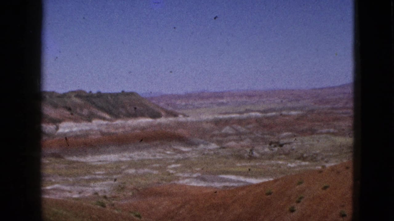 1953 California 8mm frame showing a dry red-brown landscape, pale washes, sparse scrub, and distant low hills beneath a blue sky.