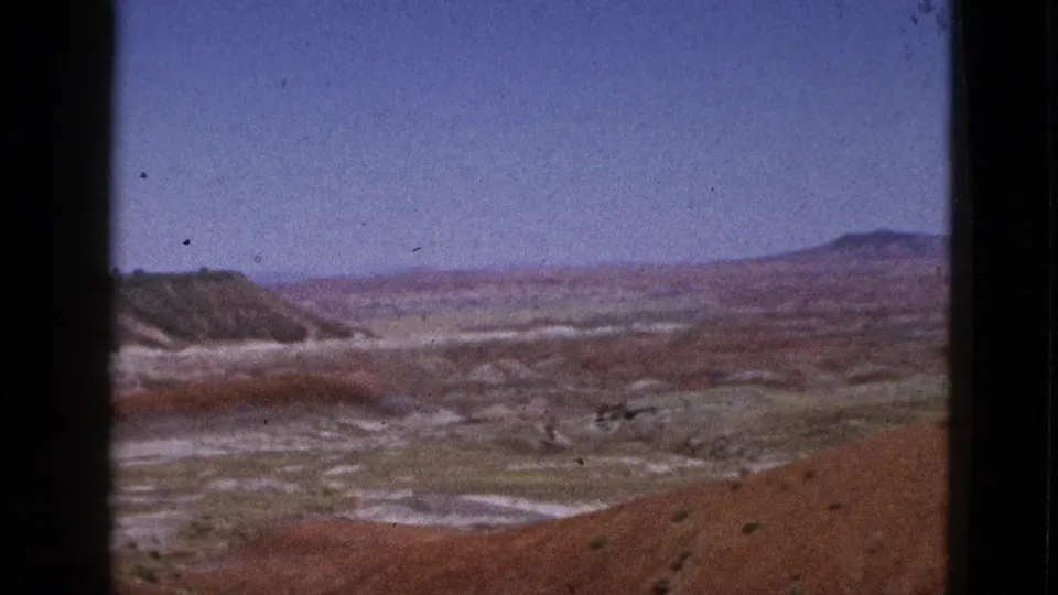 1953 California home-movie frame with a dry basin, reddish hills, pale ground patterns, sparse vegetation, and low horizon ridges.