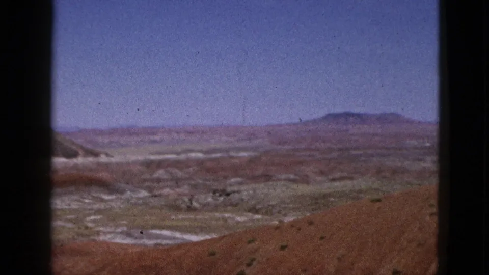 1953 California 8mm view over a red foreground hillside, pale dry washes, sparse scrub, and a distant flat-topped hill.