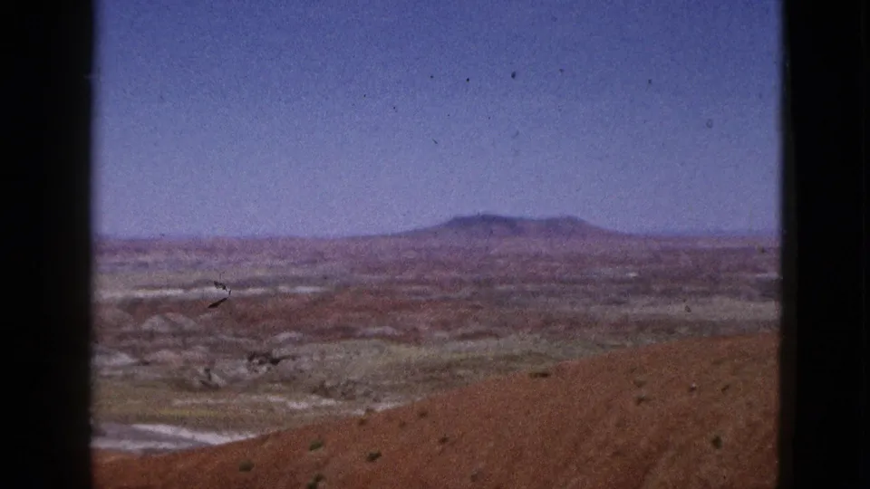 1953 California desert landscape frame showing a reddish barren slope, pale dry floor, sparse plants, and distant low hills under clear sky.