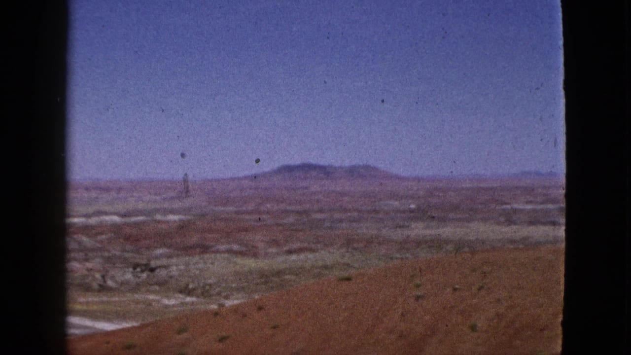 Wide desert valley with reddish foreground dirt and distant low mountains under a blue sky.