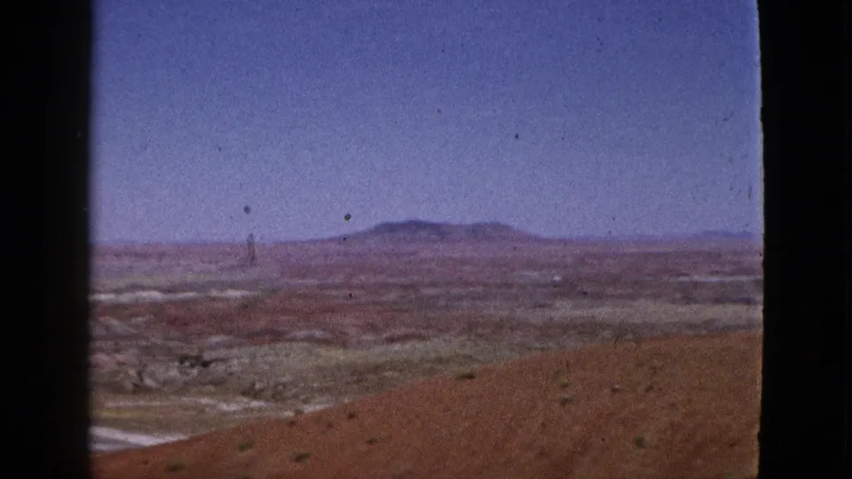 1953 California 8mm frame showing a broad desert valley, sparse foreground scrub, and hazy mountains on the horizon.
