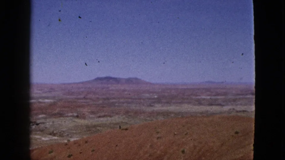 1953 California home-movie view across reddish desert ground toward a hazy valley and low mountain ridge.