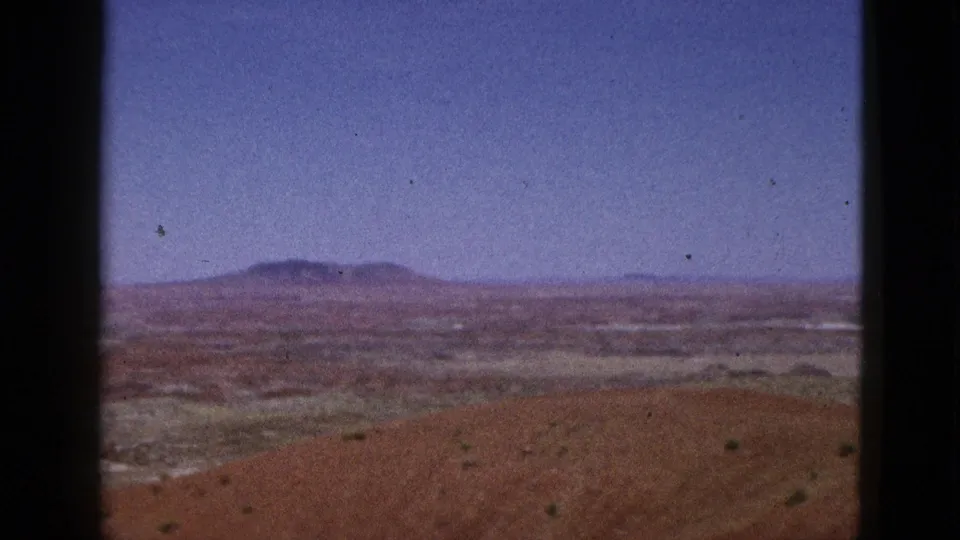 1953 California 8mm footage frame with a hazy valley floor, distant hills, and sparse plants in reddish soil.