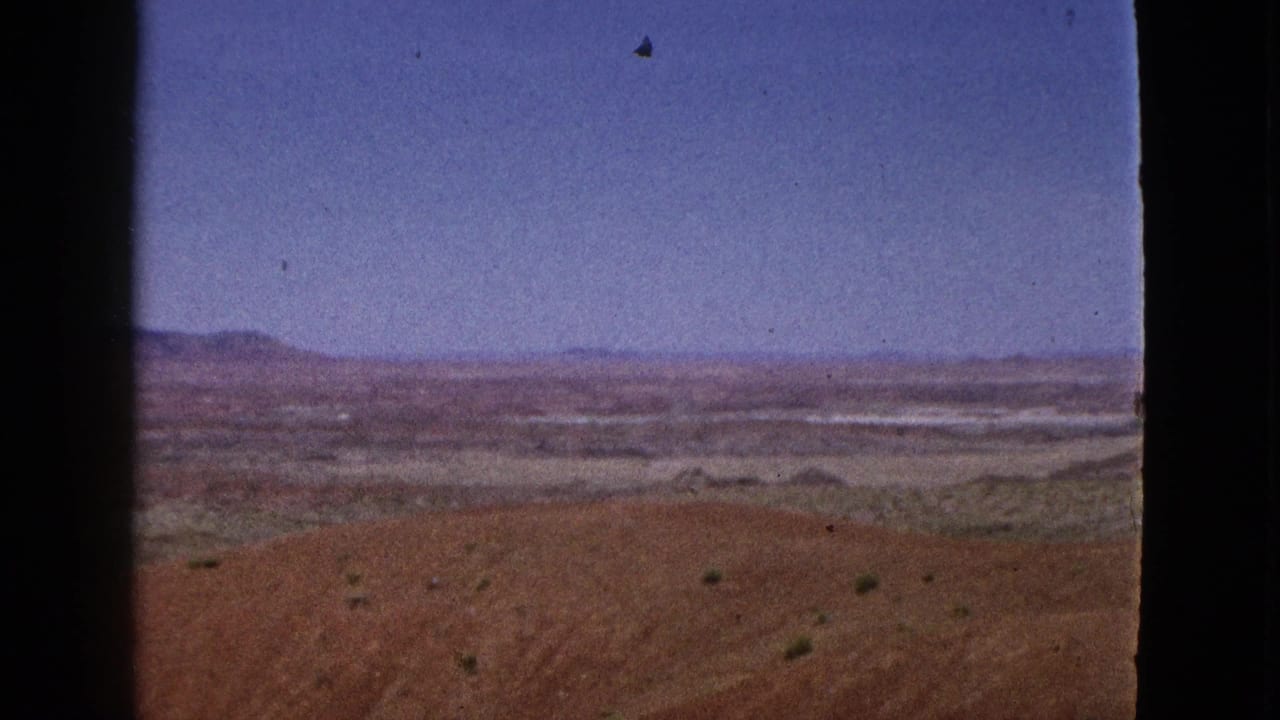 1953 California 8mm frame showing open desert terrain, low vegetation, distant hills, blue sky, and visible film borders.