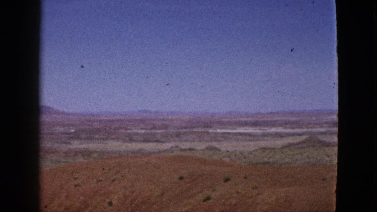 Wide desert horizon with reddish foreground, sparse vegetation, distant mountains, and blue sky divided by film border.