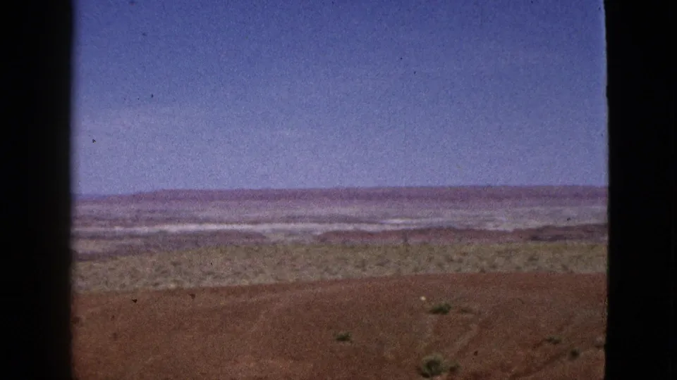 1953 California home-movie frame showing a flat desert horizon, sparse shrubs, reddish ground, blue sky, and contact-sheet film borders.