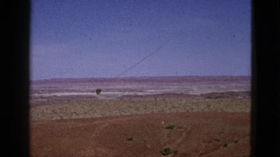 1953 California 8mm frame showing a static desert landscape with reddish soil, scattered shrubs, pale distance, and open blue sky.