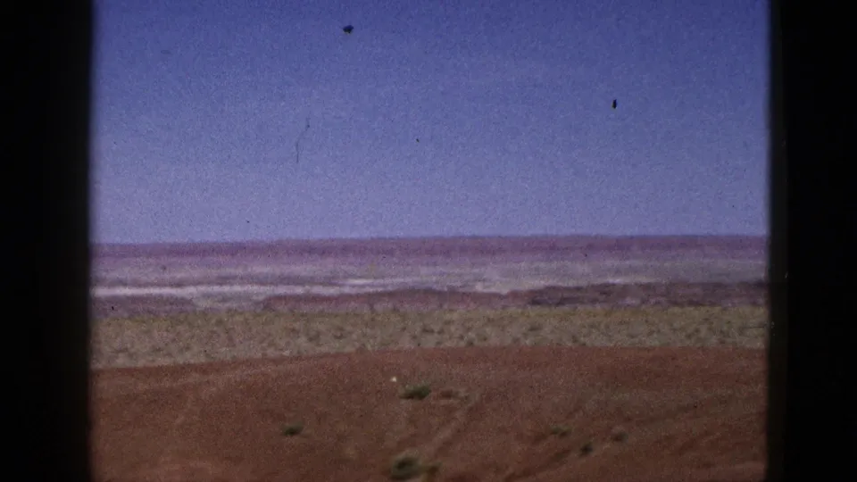 1953 California home-movie frame of open desert terrain, with red earth in front, scattered vegetation, and a broad blue sky above.