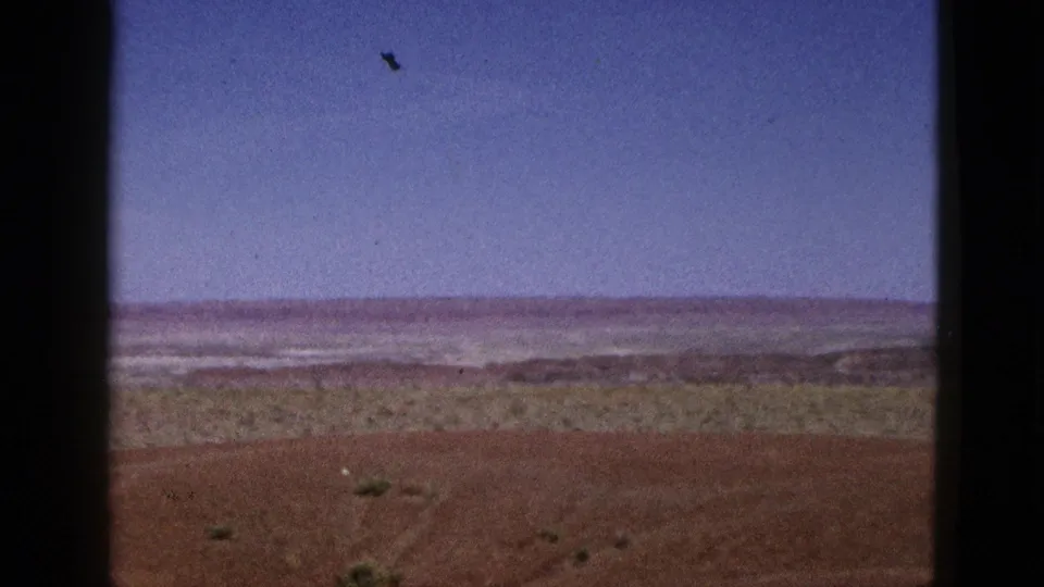 1953 California 8mm still showing a broad arid view with red soil, sparse brush, pale distant ground, and visible film dust.