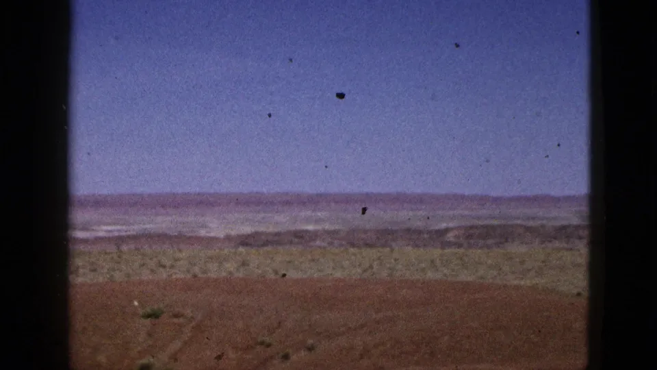 1953 California desert frame with reddish earth, low vegetation, a pale horizontal expanse, blue sky, and dark 8mm border marks.
