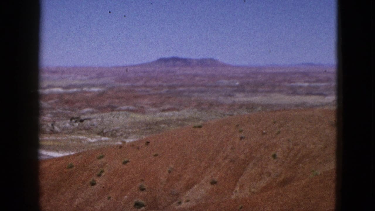 1953 California 8mm frame showing an arid red-brown slope, scattered scrub, pale desert flats, and a distant plateau beneath clear blue sky.