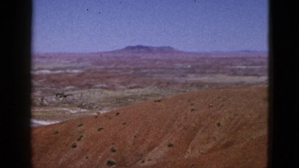1953 California 8mm frame showing an arid red-brown slope, scattered scrub, pale desert flats, and a distant plateau beneath clear blue sky.