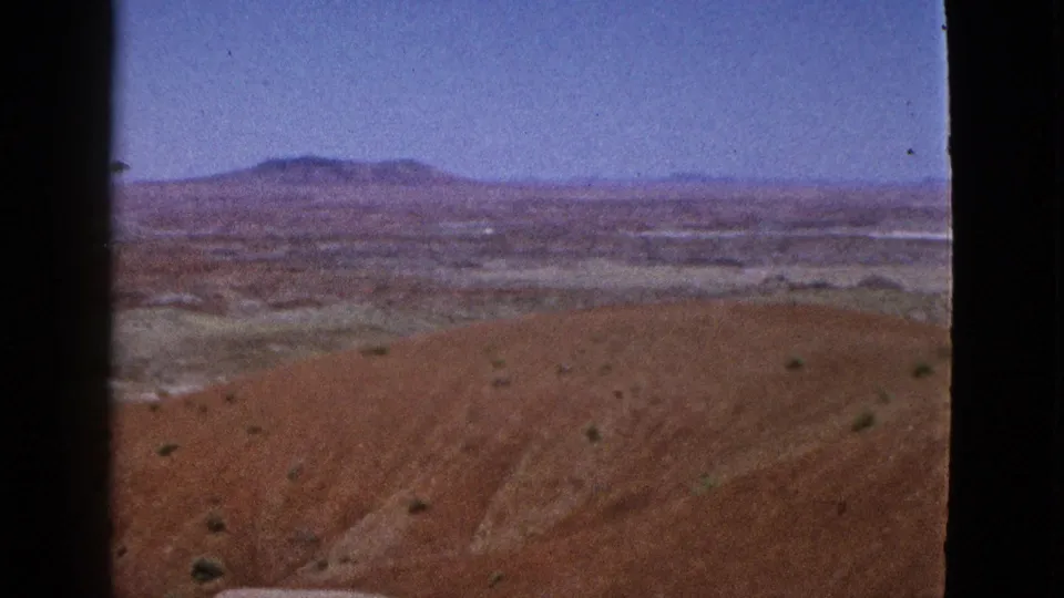 1953 California home-movie frame with reddish foreground terrain, scattered desert scrub, pale flats, and a low butte or plateau on the horizon.