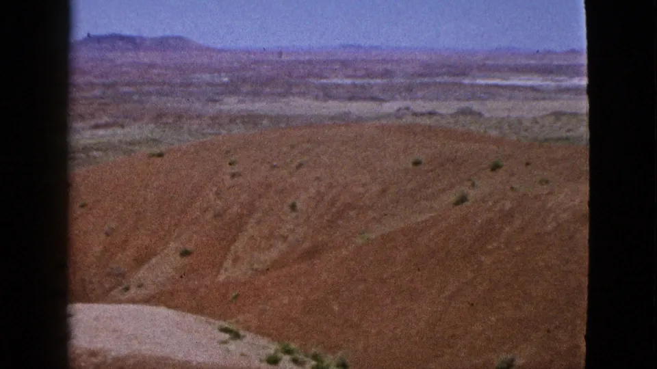 1953 California 8mm view across a red desert slope, a pale wash at the lower edge, scattered scrub, and distant flatlands fading into low ridges.