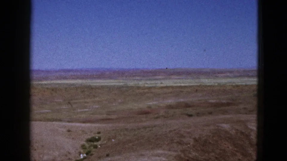 1953 California home-movie frame looking over a broad arid plain with scattered shrubs, pale flats, a distant ridge line, and clear blue sky.