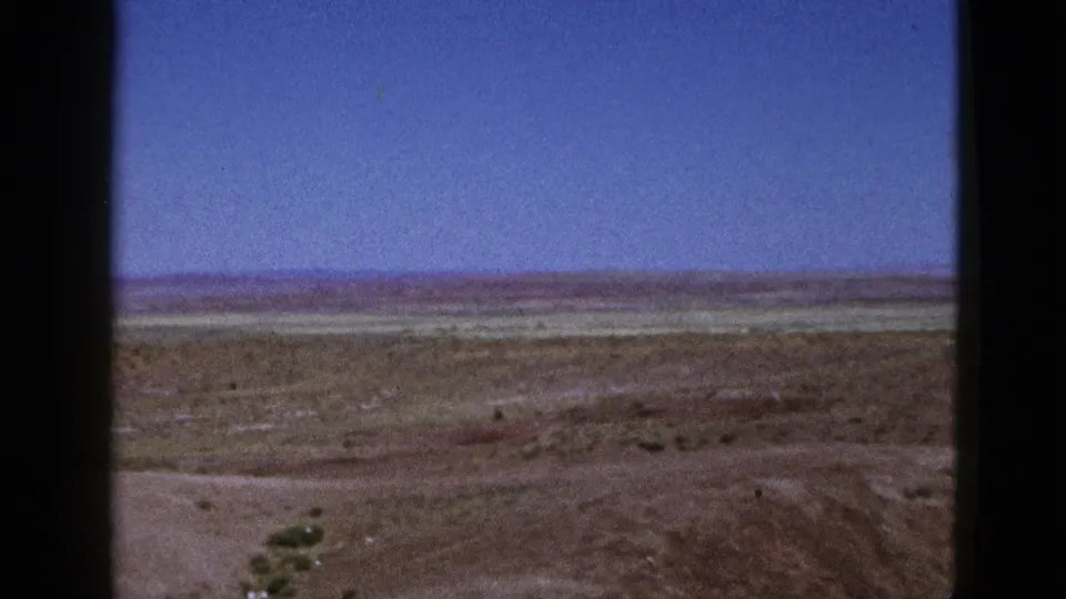 1953 California 8mm frame showing dry open ground, sparse vegetation, a distant horizon, and blue sky seen through dark framing edges.