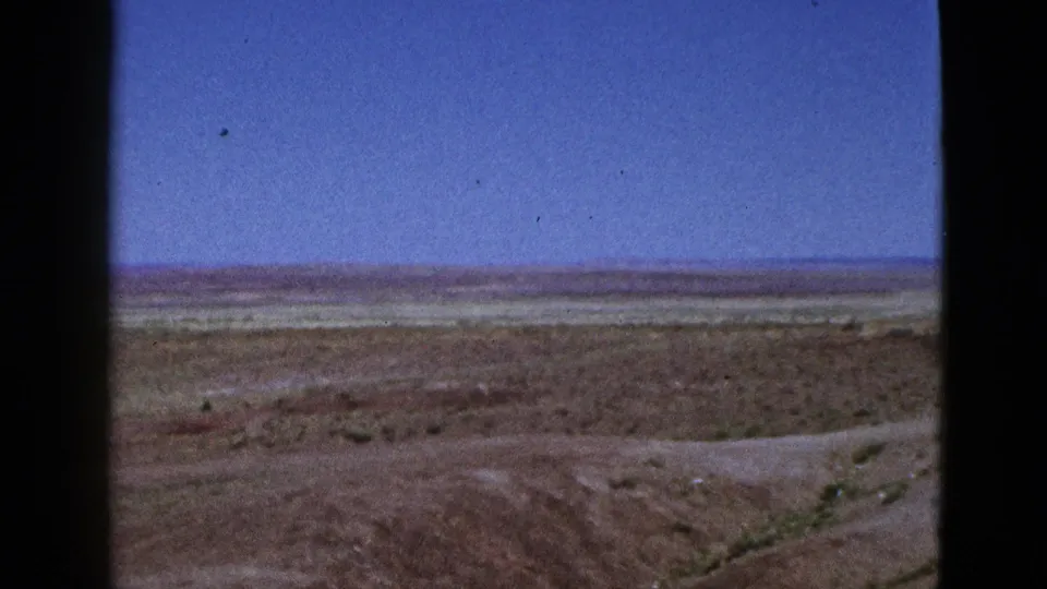 1953 California 8mm still showing a dry open field with faint ground lines, sparse plants, and a wide blue sky.