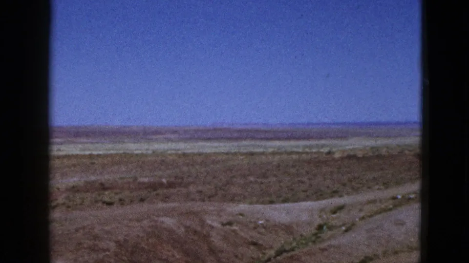 1953 California home-movie frame of an empty dry landscape with sparse vegetation, distant horizon, and clear blue sky.