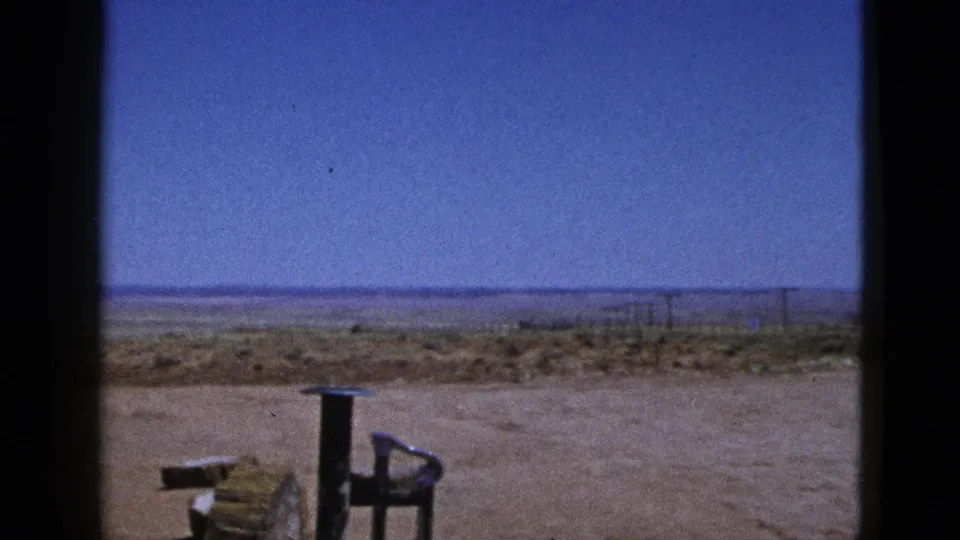 1953 California desert footage frame with a broad blue sky, pale dirt foreground, wooden object, metal frame, and distant posts.