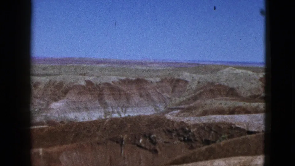 1953 California 8mm frame showing distant greenish fields, brown mesa ridges, and pale desert slopes under clear sky.