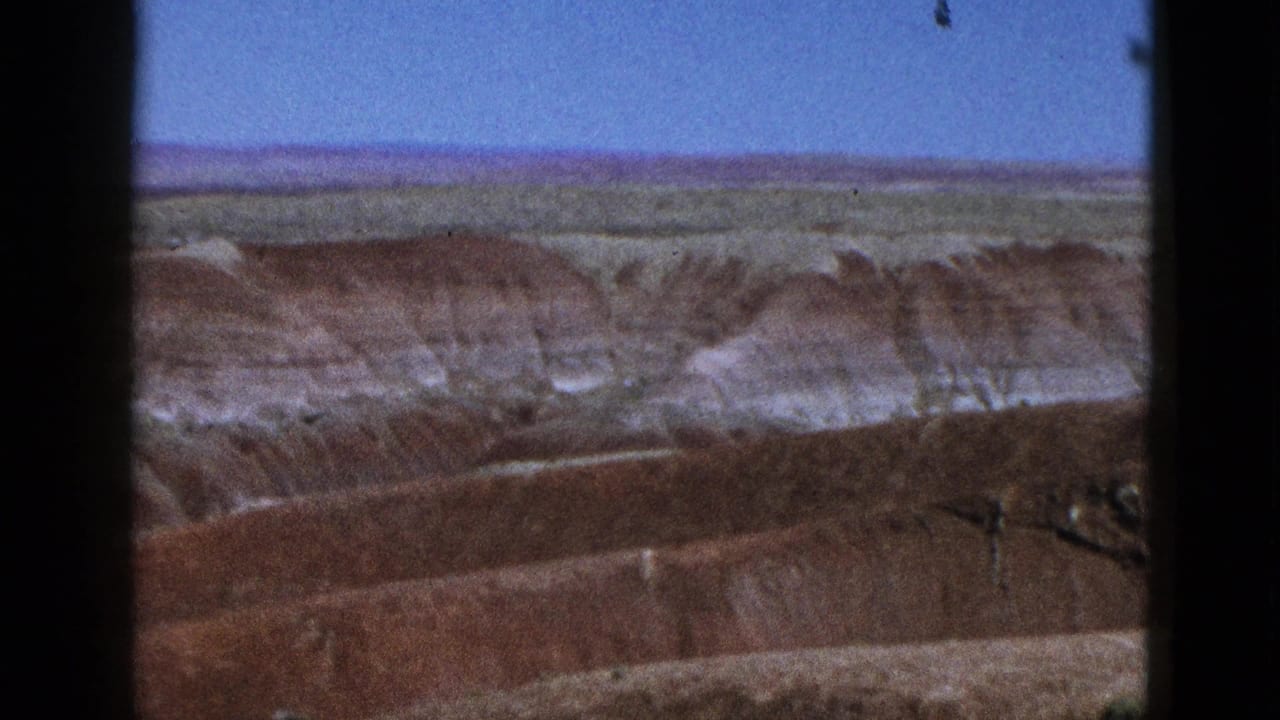 Red and white canyon ridges beneath a narrow blue sky, framed by dark film borders.
