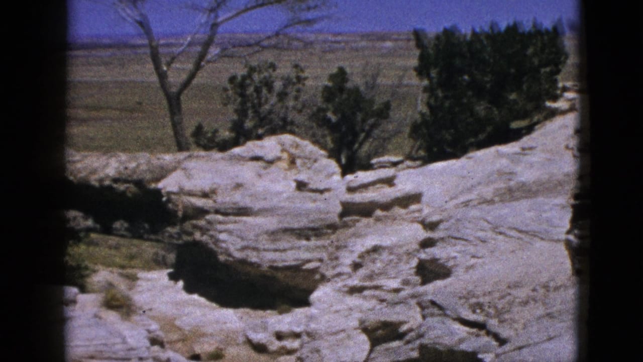 1953 California 8mm frame showing pale layered rock, scrubby trees, distant arid plains, and a saturated blue sky.