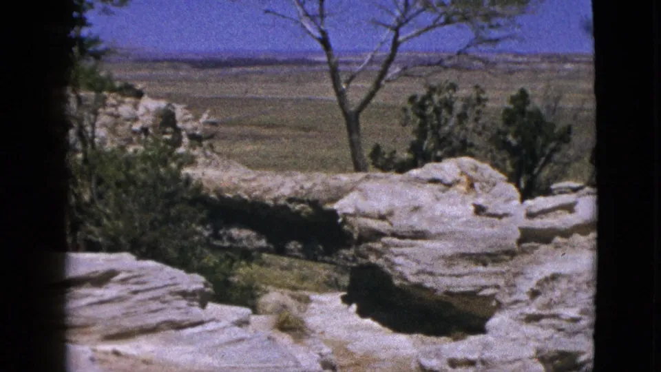 1953 California home-movie frame with a pale rock shelf in the foreground, sparse shrubs, and open plains in the distance.