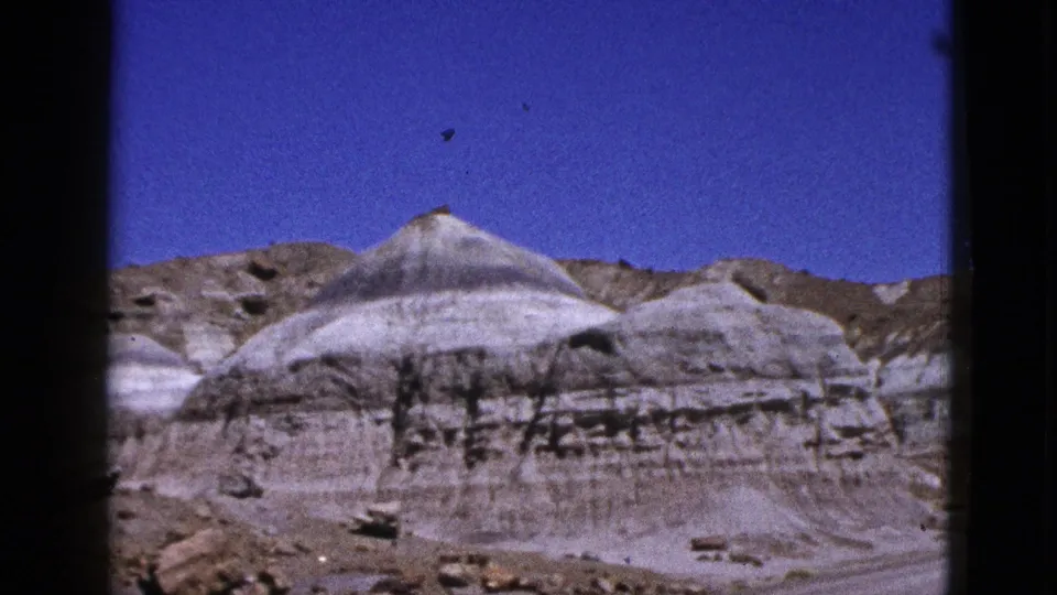 1953 California desert view with pale banded rock formations, brown surrounding hills, and a clear blue sky.