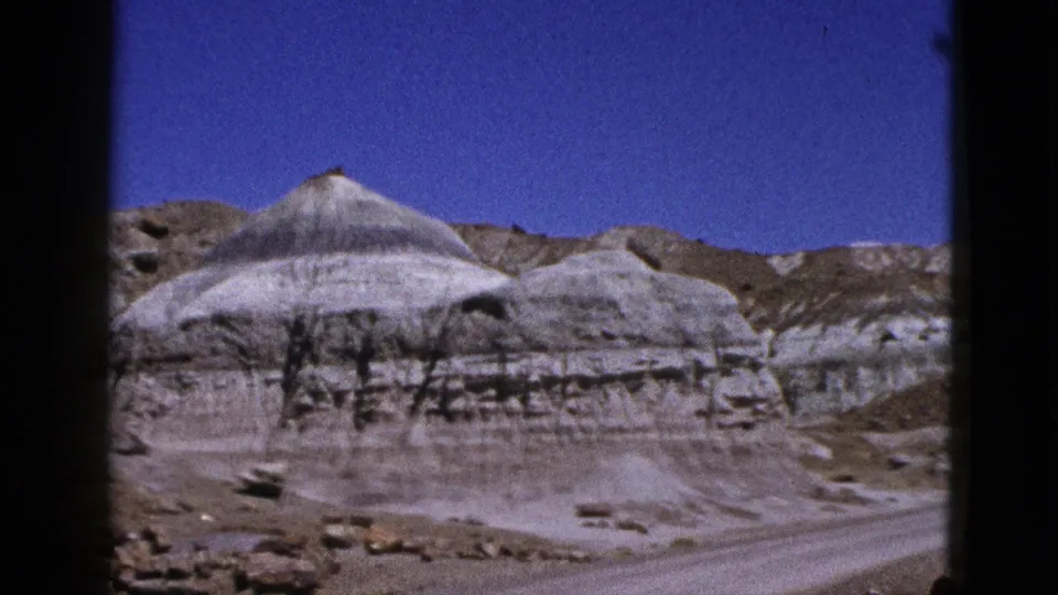 1953 California 8mm frame showing broad layered desert hills, pale eroded formations, and a dirt track across the foreground.