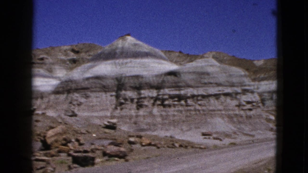 1953 California 8mm frame showing layered pale desert rock formations with a dirt road or wash below a clear blue sky.