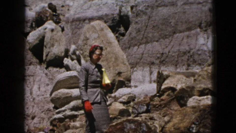 1953 California 8mm frame showing a woman in a grey dress, red gloves, and scarf standing among large desert boulders.