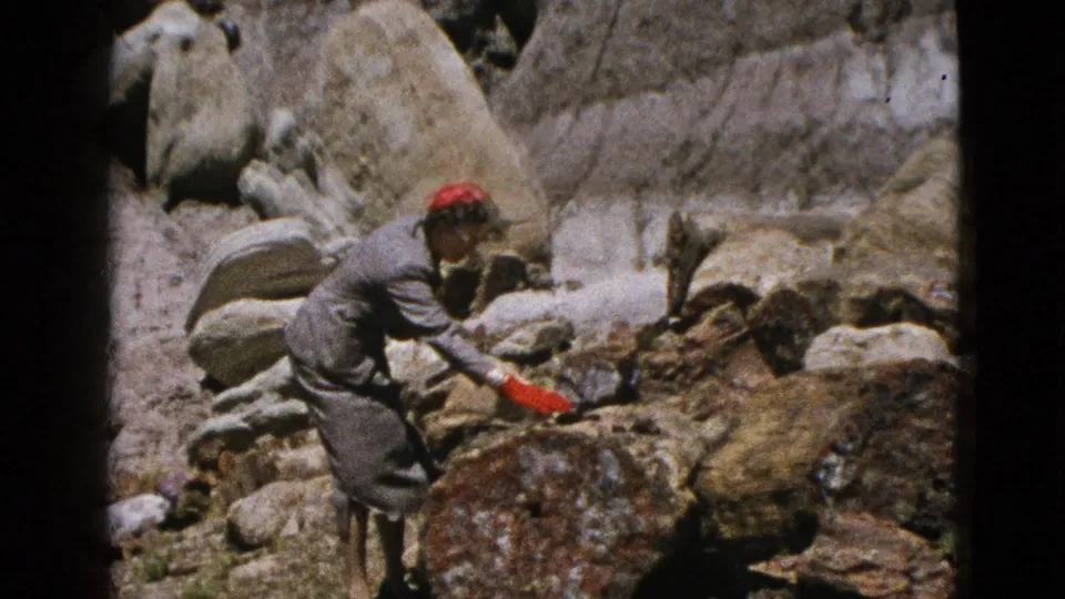 1953 California 8mm frame showing a woman in grey bending to examine or touch a large brown boulder in a rocky landscape.