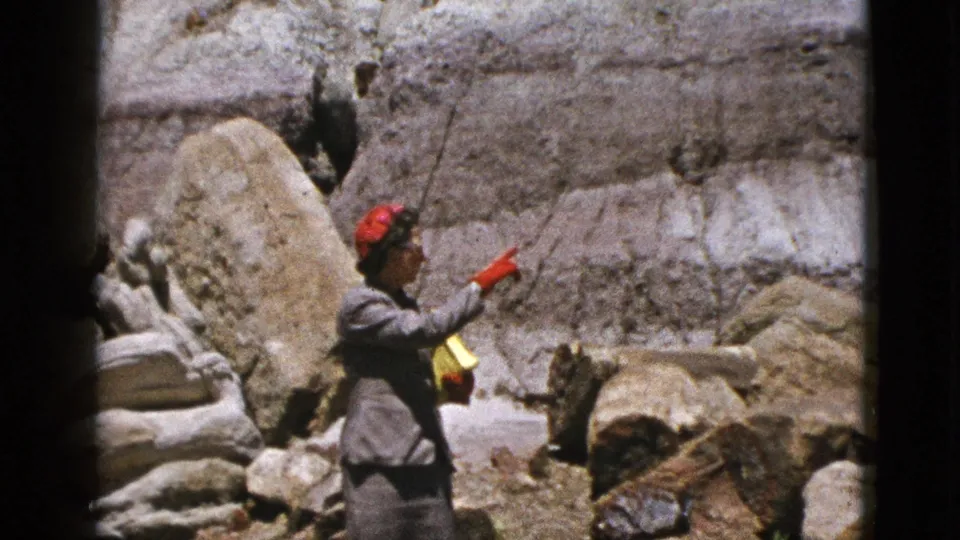 A woman in mid-century dress points toward the rocky wall while standing near petrified wood in California.