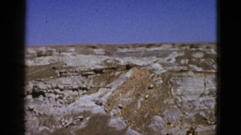 1953 California desert view with pale layered formations, rough terrain, a path-like wash, and clear blue sky in an 8mm frame.
