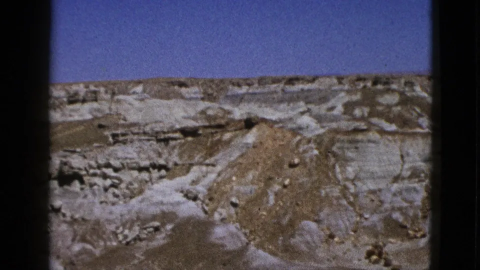 1953 California 8mm frame showing pale layered desert rock and a brown eroded mound beneath a clear blue sky.