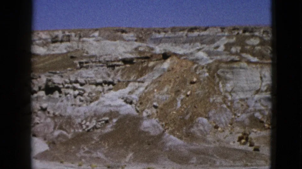 Vintage 8mm view of California desert terrain with striated rock shelves, tan mounds, and visible film-frame edges.