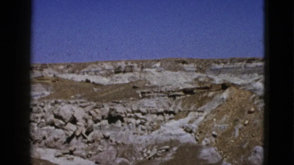 California desert landscape on 8mm film, showing pale strata, brown eroded ground, scattered rocks, and a clear sky.