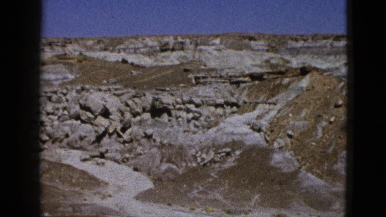 1953 Arizona 8mm frame showing an aerial view of eroded pale bluffs, darker rock bands, dry washes, and blue sky.