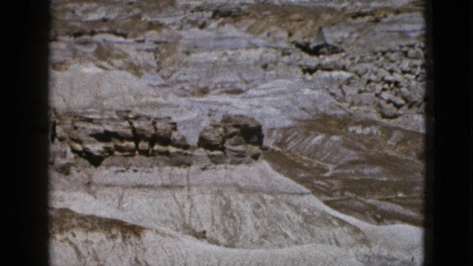 1953 Arizona 8mm aerial frame showing pale eroded formations, dark rock ledges, and dry desert channels.