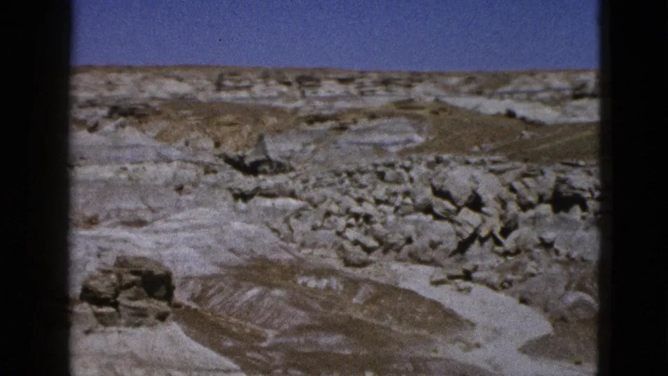 1953 Arizona 8mm frame showing pale ridges, scattered boulders, eroded slopes, and clear blue sky.