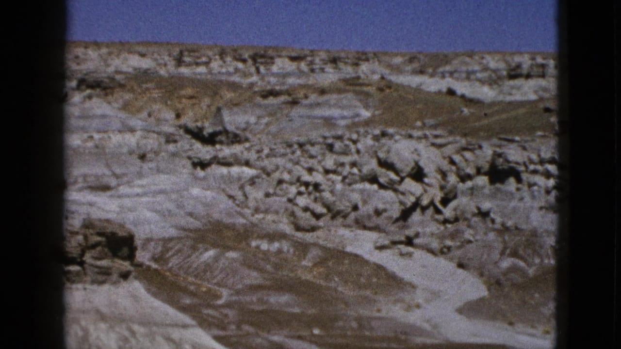 1953 Arizona home-movie frame showing eroded desert terrain, layered rock, sparse scrub, and clear blue sky.